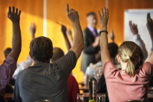 Rear view of a group of people attending a seminar or occupational training, all raising hands.